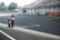 A motorcyclist is racing on an empty race track. The rider is leaning forward, wearing full gear, and appears to be speeding on a high-performance sportbike. The track is wide with a backdrop of grandstands and a tree line in the distance.