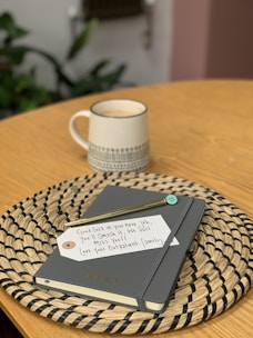 A neatly arranged scene with a ceramic mug filled with a hot beverage on a wooden table. Beside the mug is a woven placemat holding a closed journal or planner labeled 'Becky'. Resting on the planner is a gold pen and a note with a handwritten message offering good luck in a new job and expressing that the person will be missed by their outpatient family.