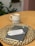 A neatly arranged scene with a ceramic mug filled with a hot beverage on a wooden table. Beside the mug is a woven placemat holding a closed journal or planner labeled 'Becky'. Resting on the planner is a gold pen and a note with a handwritten message offering good luck in a new job and expressing that the person will be missed by their outpatient family.