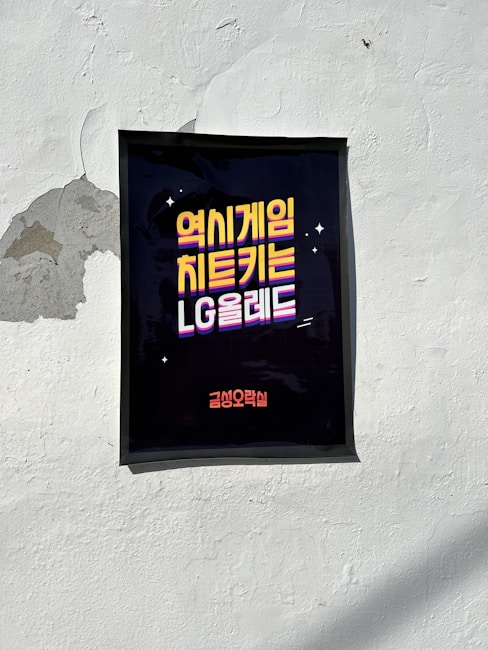 A poster with Korean text is affixed to a white, slightly weathered wall. The text is styled in bold, colorful lettering with a dark background, making it stand out prominently. The wall shows signs of wear, with peeling paint in some areas.