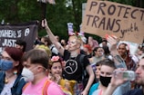 A diverse group of people are gathered for a demonstration, holding signs supporting transgender youth. Some individuals are wearing masks, and several have flowers in their hair. The scene is lively and filled with colorful attire.