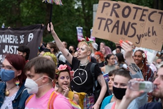 A diverse group of people are gathered for a demonstration, holding signs supporting transgender youth. Some individuals are wearing masks, and several have flowers in their hair. The scene is lively and filled with colorful attire.