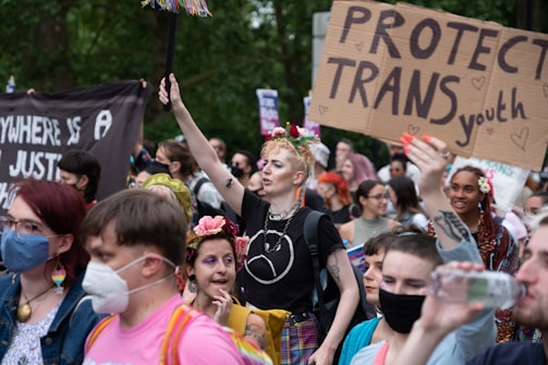 A diverse group of people are gathered for a demonstration, holding signs supporting transgender youth. Some individuals are wearing masks, and several have flowers in their hair. The scene is lively and filled with colorful attire.