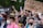 A diverse group of people are gathered for a demonstration, holding signs supporting transgender youth. Some individuals are wearing masks, and several have flowers in their hair. The scene is lively and filled with colorful attire.