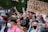 A diverse group of people are gathered for a demonstration, holding signs supporting transgender youth. Some individuals are wearing masks, and several have flowers in their hair. The scene is lively and filled with colorful attire.