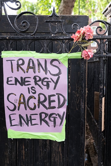 A hand-painted sign is taped to a black wrought iron fence with green tape. The sign reads 'TRANS ENERGY IS SACRED ENERGY' in black bold letters on a purple background. A bouquet of pink flowers is attached to the fence next to the sign. Green trees and a brick building can be seen in the background.