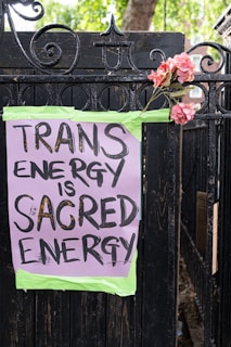 A hand-painted sign is taped to a black wrought iron fence with green tape. The sign reads 'TRANS ENERGY IS SACRED ENERGY' in black bold letters on a purple background. A bouquet of pink flowers is attached to the fence next to the sign. Green trees and a brick building can be seen in the background.