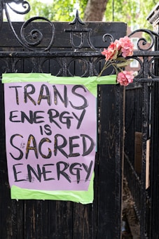 A hand-painted sign is taped to a black wrought iron fence with green tape. The sign reads 'TRANS ENERGY IS SACRED ENERGY' in black bold letters on a purple background. A bouquet of pink flowers is attached to the fence next to the sign. Green trees and a brick building can be seen in the background.