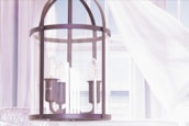 A modern light fixture with multiple bulbs encased in a cylindrical glass cover, hanging in a bright, airy room. White curtains gently billow in the background, allowing a view of a serene seascape outside the window.