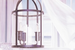 A modern light fixture with multiple bulbs encased in a cylindrical glass cover, hanging in a bright, airy room. White curtains gently billow in the background, allowing a view of a serene seascape outside the window.