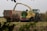 A large agricultural machine is actively harvesting a field, transferring cut crops into an adjacent container truck. The foreground features some foliage, partially framing the scene. The landscape is flat, with a gray overcast sky in the background.