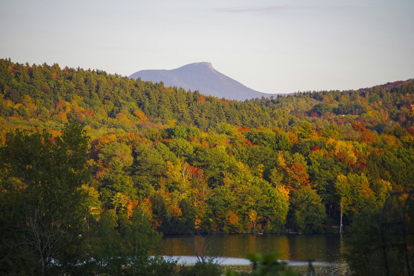 The distinctive humped summit of a mountain rising above rolling forested hills in early autumn color, with a lake in the foreground