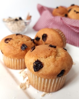 Three golden-brown muffins with dark raisins are placed on a white surface. In the background, there's a bowl of oats and a basket with more muffins, lined with a red and white checkered cloth. Crumbs are scattered around the muffins.