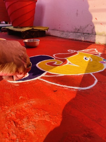 A person's hand is creating a colorful art design on the floor using powdered pigments. The design appears to be a traditional rangoli pattern with vivid colors such as yellow and purple against a red background. A silver bowl is nearby, and a red plant pot is visible in the background.