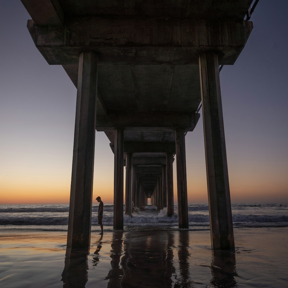 a person standing under a pier at sunset