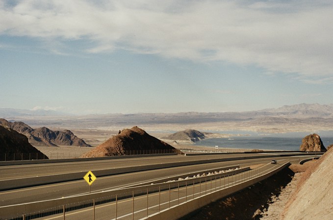 A desert highway running toward rocky mountains and open water, similar to the terrain around Boulder City and the Eldorado Valley