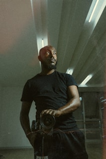 A person wearing a black shirt stands in a room with modern, minimalistic architecture. The lighting casts a dramatic shadow, highlighting the person's contemplative expression as they look away from the camera, holding a camera in one hand. The ceiling has regular patterns with built-in lights, contributing to the ambience.