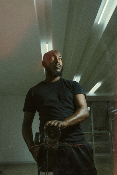 A person wearing a black shirt stands in a room with modern, minimalistic architecture. The lighting casts a dramatic shadow, highlighting the person's contemplative expression as they look away from the camera, holding a camera in one hand. The ceiling has regular patterns with built-in lights, contributing to the ambience.