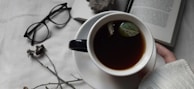 A cup of dark-colored tea with floating leaves held by a person in a white sweater on a white cloth surface. Nearby are scattered dried flowers, a pair of black glasses, and an open book.