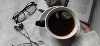 A cup of dark-colored tea with floating leaves held by a person in a white sweater on a white cloth surface. Nearby are scattered dried flowers, a pair of black glasses, and an open book.