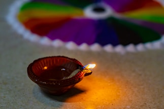 A small clay oil lamp with a visible flame is placed on a surface, creating a warm glow around it. In the background, blurred, there is a rangoli design made with vibrant colors including green, red, purple, and yellow, possibly with a radial pattern.