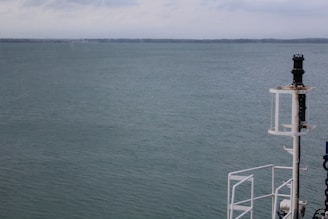 A calm ocean with serene water stretching out to the horizon under a slightly overcast sky. On the right side of the image is a part of a ship's structure, including a navigation light or equipment post.