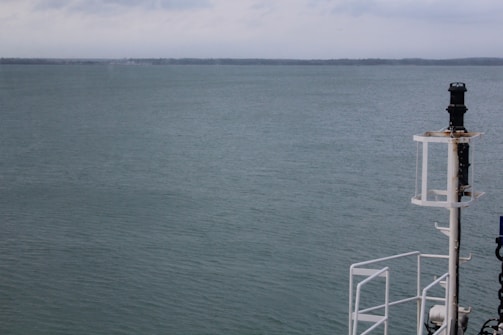 A calm ocean with serene water stretching out to the horizon under a slightly overcast sky. On the right side of the image is a part of a ship's structure, including a navigation light or equipment post.