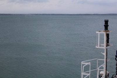 A calm ocean with serene water stretching out to the horizon under a slightly overcast sky. On the right side of the image is a part of a ship's structure, including a navigation light or equipment post.