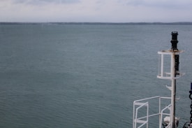 A calm ocean with serene water stretching out to the horizon under a slightly overcast sky. On the right side of the image is a part of a ship's structure, including a navigation light or equipment post.