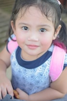 A young child with dark hair and expressive eyes looks directly at the camera. The child wears a patterned shirt with floral designs and a pink backpack. The background is softly blurred, emphasizing the child's face and expression.