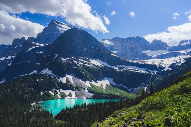 A turquoise alpine lake ringed by pines below dark snow-streaked peaks and a blue summer sky