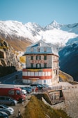 A historic hotel building with red and white paint stands adjacent to a winding mountain road, surrounded by parked vehicles and several people. Snow-capped mountains rise in the background, contrasting with the clear blue sky.