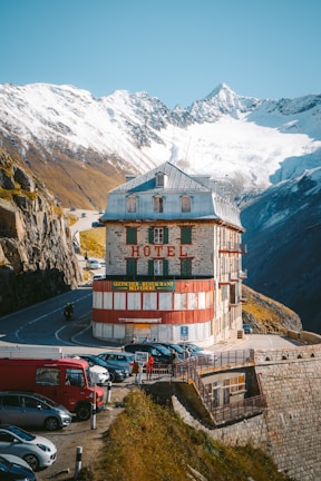 A historic hotel building with red and white paint stands adjacent to a winding mountain road, surrounded by parked vehicles and several people. Snow-capped mountains rise in the background, contrasting with the clear blue sky.