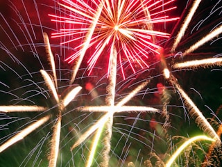 A vibrant display of fireworks with bright red and white streaks against a dark sky. The explosive patterns create star-like shapes that illuminate the surroundings, showcasing an elegant cascade of light.