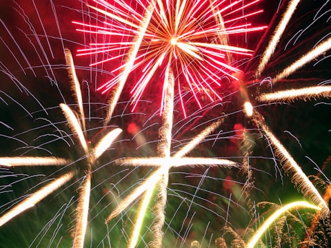 A vibrant display of fireworks with bright red and white streaks against a dark sky. The explosive patterns create star-like shapes that illuminate the surroundings, showcasing an elegant cascade of light.