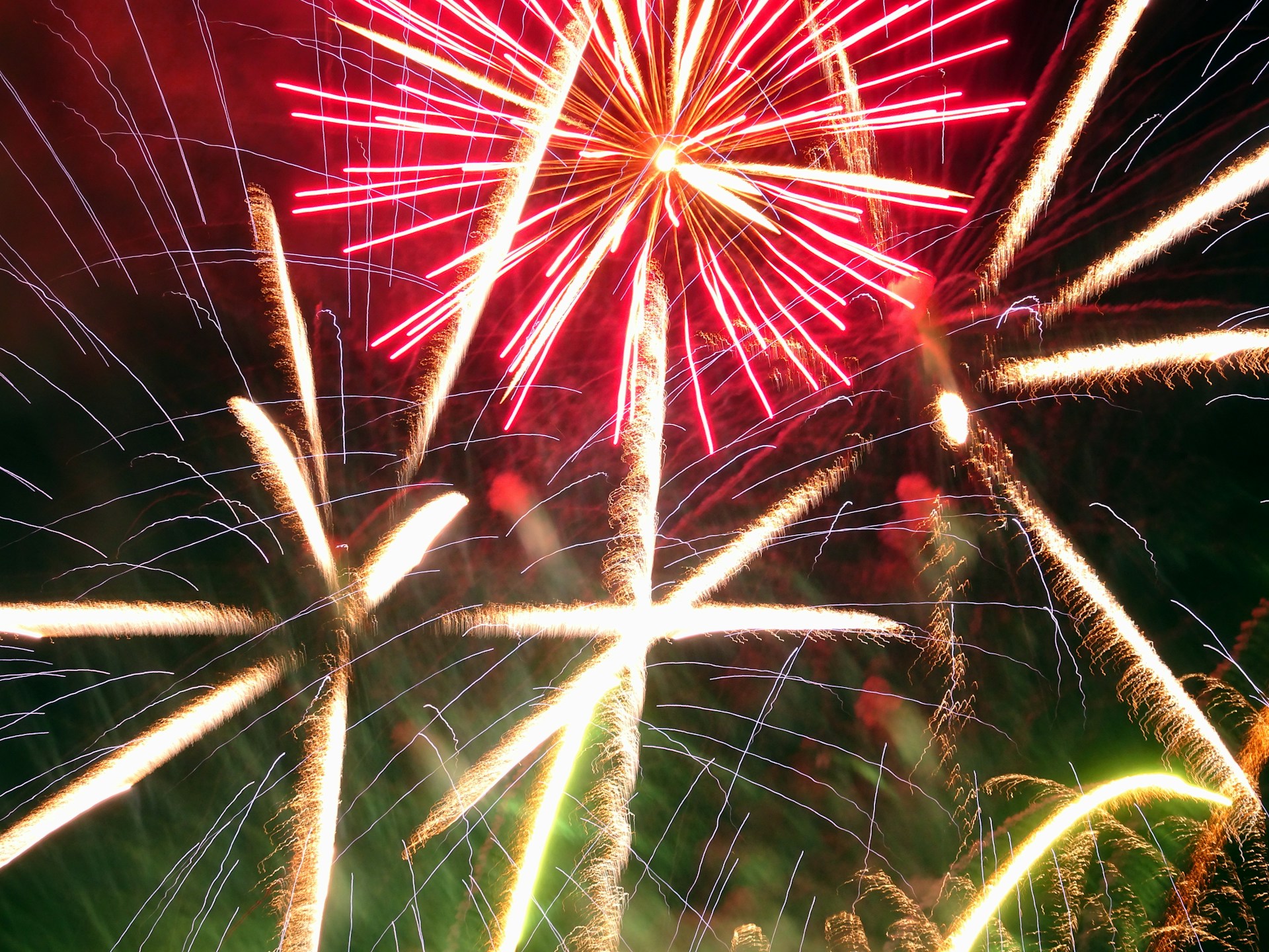 A vibrant display of fireworks with bright red and white streaks against a dark sky. The explosive patterns create star-like shapes that illuminate the surroundings, showcasing an elegant cascade of light.