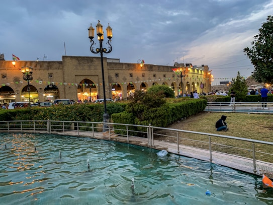 A vibrant public space with a classical brick building illuminated by lights in the background. The scene includes a small park area with trimmed hedges and a pool of water surrounded by a railing in the foreground. People are casually walking or sitting in the park, and there are decorations and flags hanging above the building.