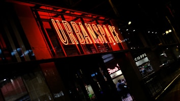 A brightly illuminated neon sign reading 'URBANSPACE' in vivid red dominates the scene. Below it, an interior view of a caf&eacute; or similar establishment features visible seats and people inside. The overall setting appears urban and bustling, capturing a nighttime atmosphere.