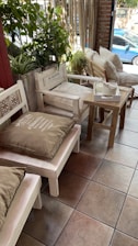 A cozy indoor seating area with wooden furniture, including benches and chairs adorned with large cushions. There are green plants in the background, adding a touch of natural decor. A small wooden table holds a tissue box, and sunlight is gently streaming in through a large window, in front of which a blue car is visible outside.