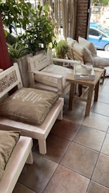 A cozy indoor seating area with wooden furniture, including benches and chairs adorned with large cushions. There are green plants in the background, adding a touch of natural decor. A small wooden table holds a tissue box, and sunlight is gently streaming in through a large window, in front of which a blue car is visible outside.