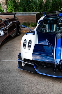 A close-up view of two luxury sports cars parked outdoors. The car in the foreground features a sleek white and blue design with intricate details such as carbon fiber elements and distinctive headlights. The second car, partially visible, has a dark exterior with a sporty appearance.