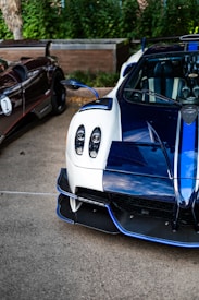 A close-up view of two luxury sports cars parked outdoors. The car in the foreground features a sleek white and blue design with intricate details such as carbon fiber elements and distinctive headlights. The second car, partially visible, has a dark exterior with a sporty appearance.
