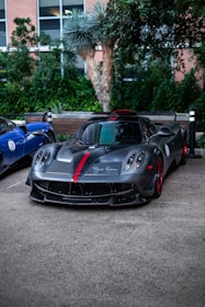 A sleek, high-performance sports car with a matte black finish and red accents is parked on a concrete surface. Lush greenery and a building with large windows form the background, adding a touch of nature to the urban setting. The car's aerodynamic design and styled headlights are distinctive features.