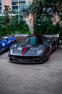 A sleek, high-performance sports car with a matte black finish and red accents is parked on a concrete surface. Lush greenery and a building with large windows form the background, adding a touch of nature to the urban setting. The car's aerodynamic design and styled headlights are distinctive features.