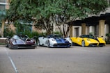 Three high-performance sports cars are parked side by side in an outdoor setting with trees and a building in the background. The cars feature sleek designs and vibrant colors, including black and red, white and blue, and yellow and black.
