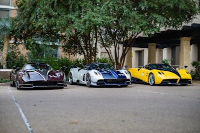Three high-performance sports cars are parked side by side in an outdoor setting with trees and a building in the background. The cars feature sleek designs and vibrant colors, including black and red, white and blue, and yellow and black.