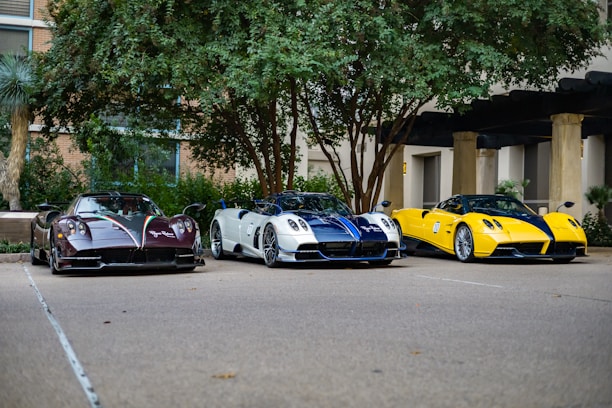 Three high-performance sports cars are parked side by side in an outdoor setting with trees and a building in the background. The cars feature sleek designs and vibrant colors, including black and red, white and blue, and yellow and black.