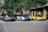 Three high-performance sports cars are parked side by side in an outdoor setting with trees and a building in the background. The cars feature sleek designs and vibrant colors, including black and red, white and blue, and yellow and black.