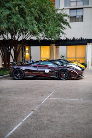 Several high-end sports cars are parked side by side in front of a building with large windows. The cars display sleek, aerodynamic designs with vibrant colors. A lush green tree hangs over the cars, and the building facade provides a contrasting urban backdrop.