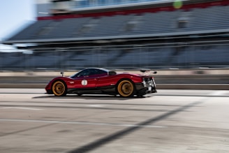 A high-performance red sports car with golden rims is speeding past a grandstand on a racetrack. The background is slightly blurred, emphasizing the motion and speed of the car. The grandstand appears empty, with multiple rows of gray seating under a partly cloudy sky.